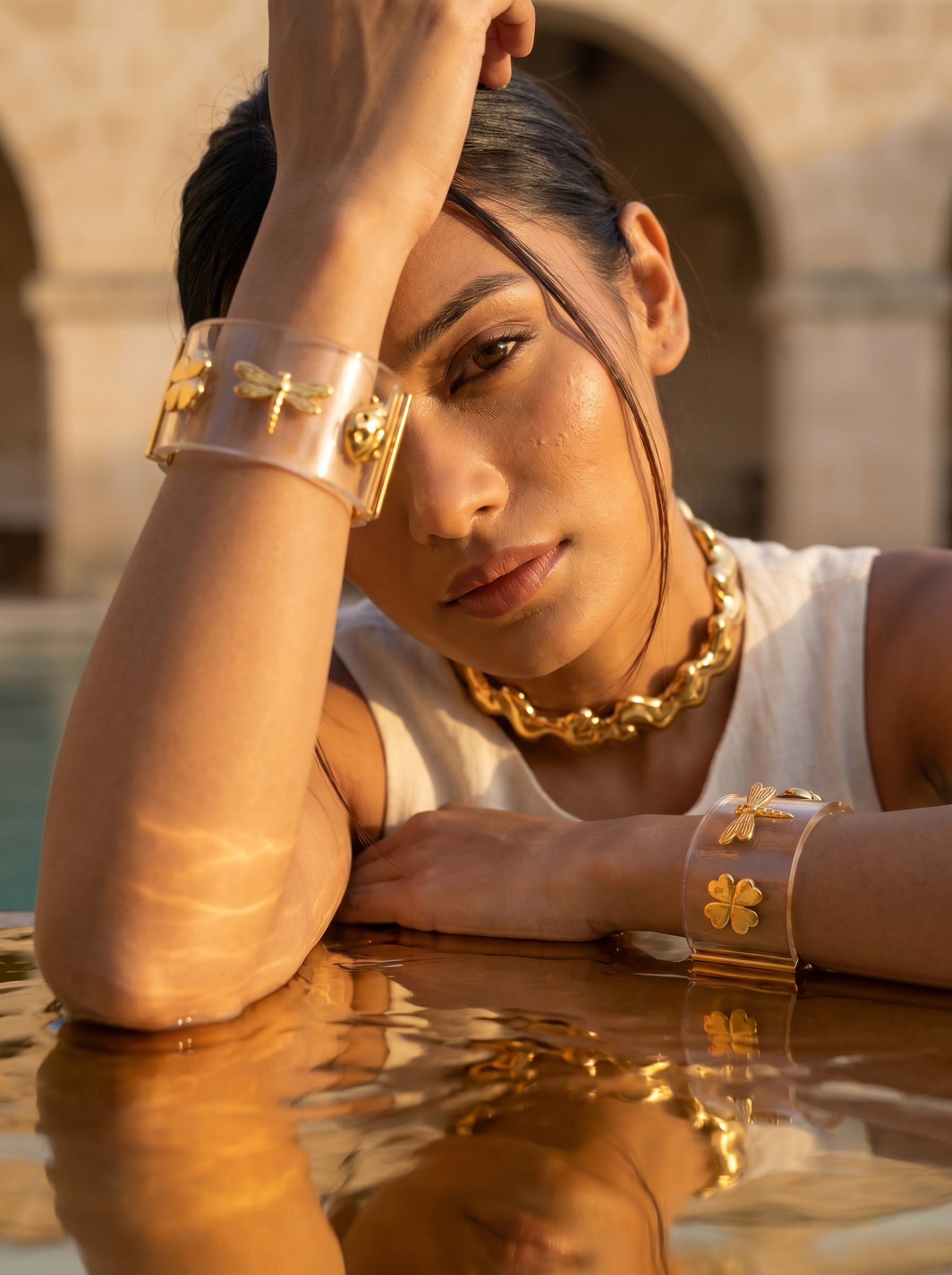 A woman resting her arms at the water’s edge wearing a chunky gold chain necklace and clear cuffs set with gold charms